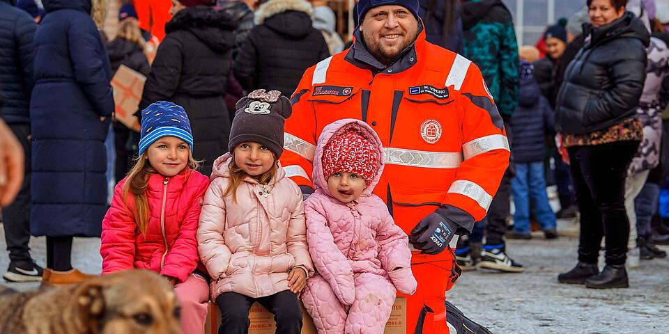 Johanniter steht hinter drei Kindern, die lächelnd auf einem Weihnachtstruckerpaket sitzen