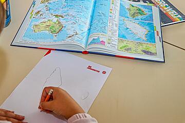 Ein Kind zeichnet mit einem roten Stift auf einem Blatt Papier, daneben liegt ein aufgeschlagener Atlas.