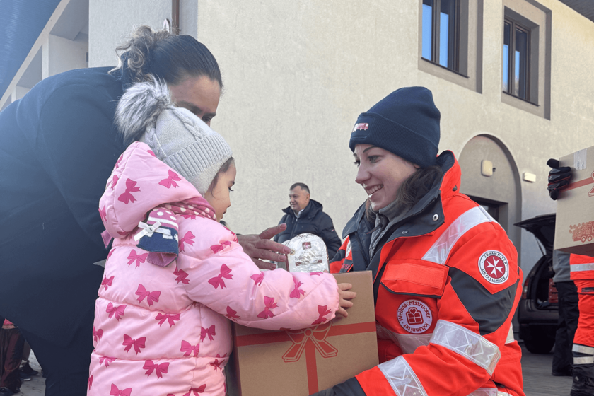 Eine Johanniter-Helferin in orangeroter Einsatzkleidung übergibt draußen ein Paket mit Geschenkaufdruck an ein kleines Mädchen im rosafarbenen Wintermantel. 