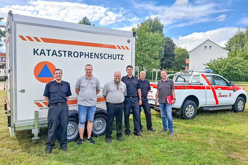 Sechs Personen stehen vor dem Kühlanhänger und einem Transportfahrzeug der Johanniter.
