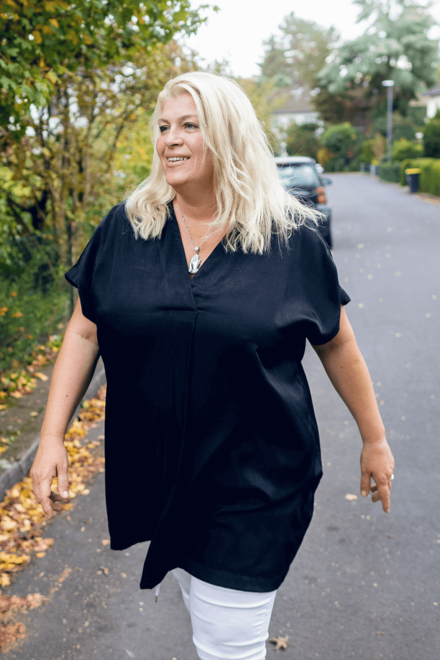 Blonde Frau in einem dunkelblauen Oberteil und offenen Haaren. Hinter ihr eine Nebenstraße mit Büschen, Hecken, einem Auto und Eingängen zu Vorgärten.