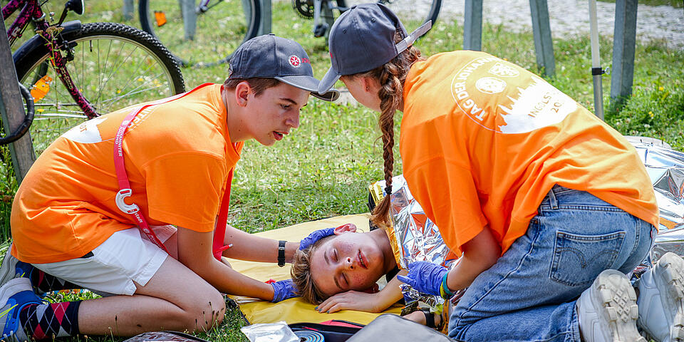 Zwei Ersthelfer in Orange versorgen bewusstlose Person mit Rettungsdecke im Gras.
