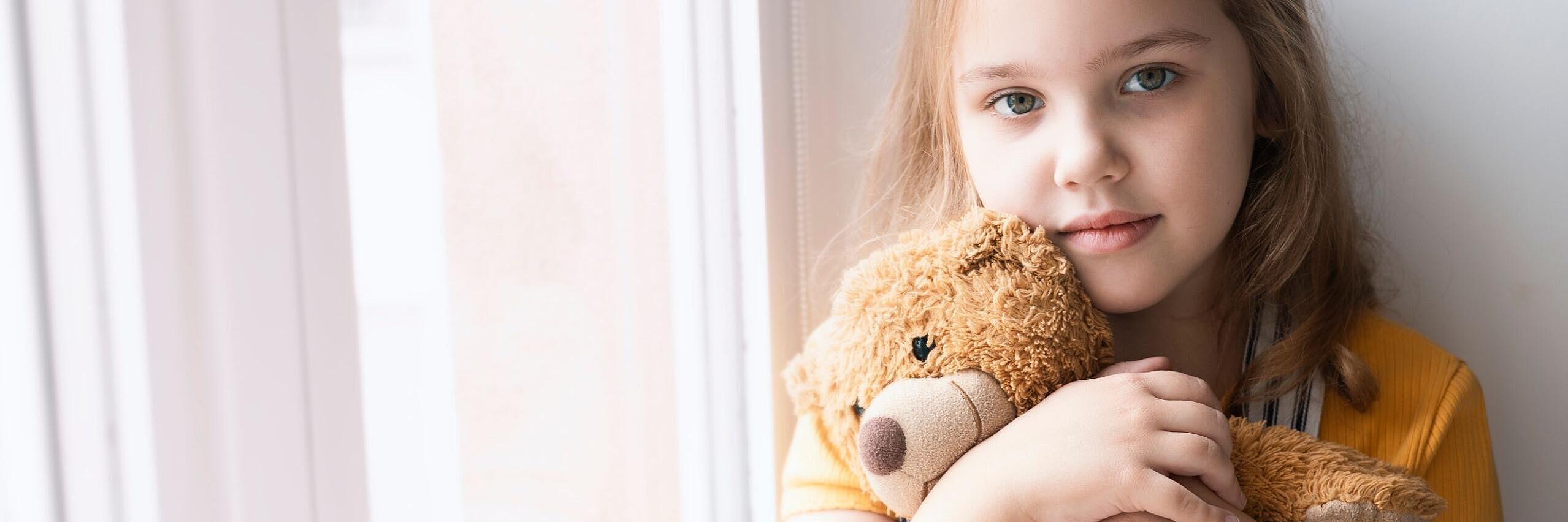 Ein Mädchen steht an einem Fenster und hält einen Teddybär im Arm.