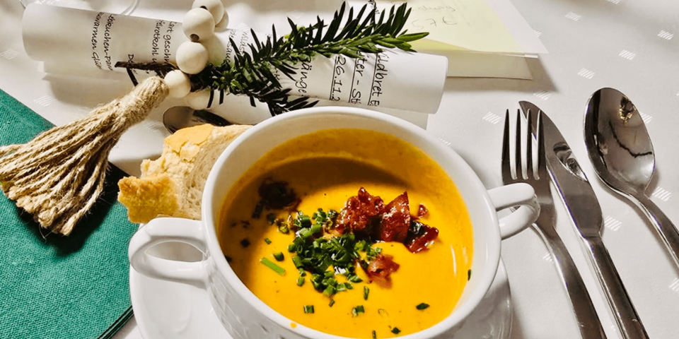 Teller mit cremiger Suppe, garniert mit Kräutern und Speck, dazu ein Stück Brot, Besteck und weihnachtliche Tischdeko