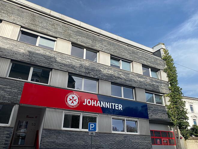 Zu sehen ist ein vierstöckiges Gebäude mit einem großen Schild, auf dem Johanniter steht. Links sieht man eine Eingangstür, rechts ein Garagentor.