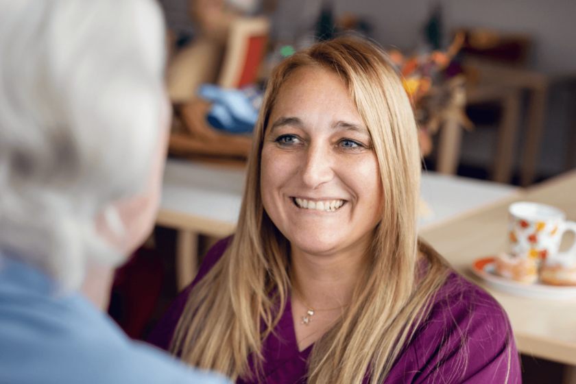 Eine Frau mit lange rötliche-blonde Haare lächelt eine Person mit grauen Haaren an, deren Hnterkopf angeschnitten und verschwommen von hinten zu sehen ist