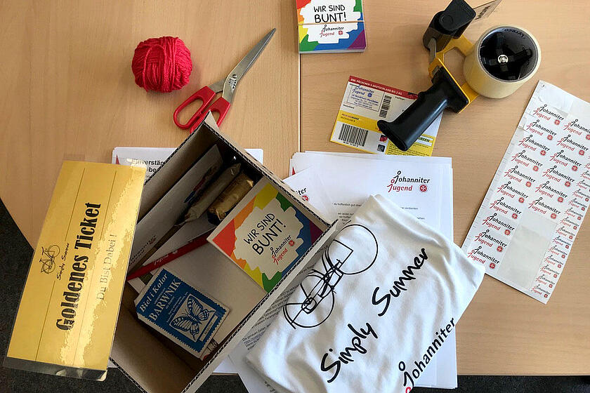 Ein Paket liegt ausgepackt auf einem Tisch. Darin sind unter anderem Aufkleber, ein T-Shirt mit Simply-Summer-Aufdruck, ein Faden, Süßigkeiten, Ein Kugelschreiber.
