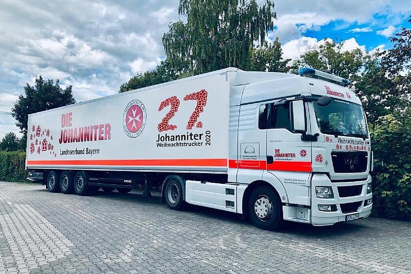 Ein großer Johanniter-Lkw mit der Aufschrift „Johanniter Weihnachtstrucker 2020“ steht auf einem Parkplatz vor Bäumen.