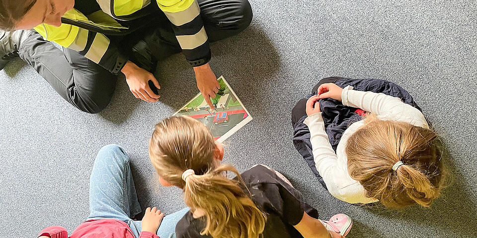 Eine Polizistin sitzt mit 3 Kindern auf einem Fußboden und zeigt ihnen ein Comicblatt