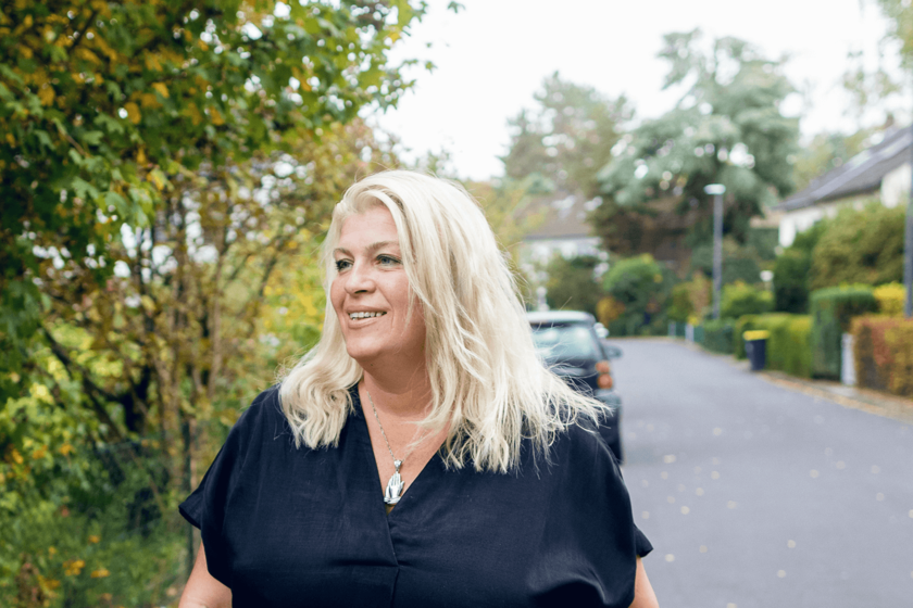 Blonde Frau in einem dunkelblauen Oberteil und offenen Haaren. Hinter ihr eine Nebensrtaße mit Büschen, Hecken, einem Auto und Eingängen zu Vorgärten.