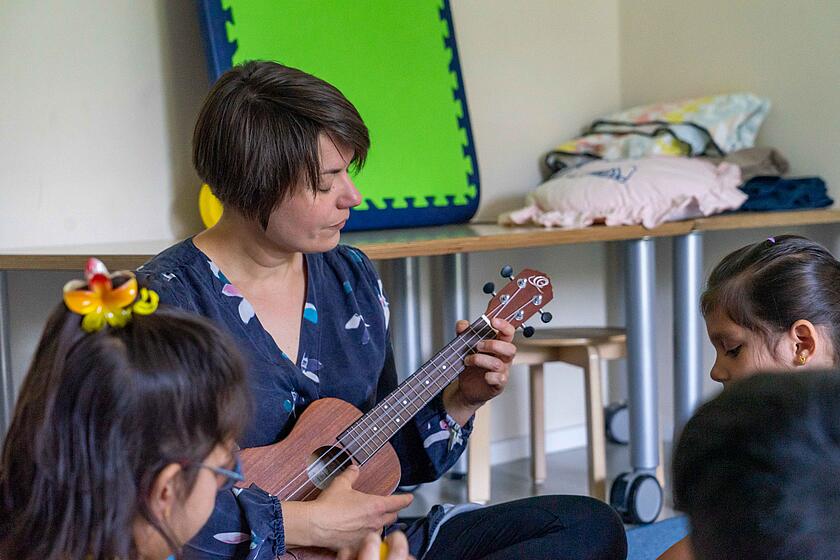 Eine Frau spielt eine Ukulele, im Vordergrunds sitzen Kinder.